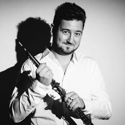 Black and white image of Philippe Tondre holding his oboe smiling whilst wearing a long sleeve white shirt